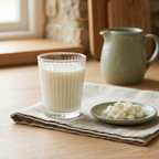 Glass of creamy milk kefir beside white kefir grains on a sage-green dish, with natural linen and a matching ceramic jug.