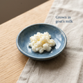 White kefir grains in a blue ceramic dish on natural linen, with the caption “Grown in goat’s milk”.