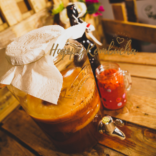 Cloth-covered Kilner tap jar filled with amber kombucha on a wooden tray beside a bottle and red patterned glass